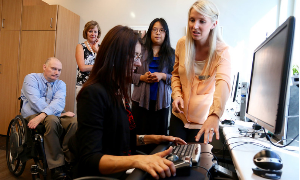 Lady in a wheel chair sitting at a computer and looking at the keyboard. There is a women assisting her.